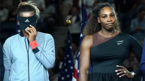 AFP Naomi Osaka pulls her visor down covering her face, Serena Williams stands hand on one hip