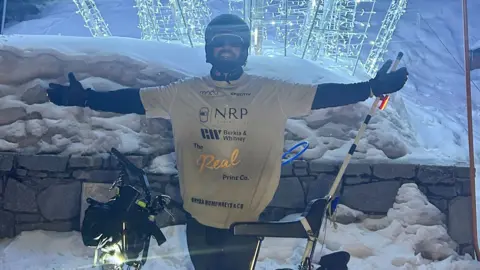 Mike Humphreys stood with his Chopper at Val Thorens in the French Alps. He is wearing a helmet and sun glasses and is standing in front of a wall and a curtain of fairy lights with his arms outstretched.

