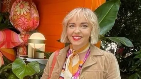 Johanna Haughey smiling at the camera.  She hair bobbed blonde hair and is wearing red lipstick, a beige jacket with fringed sleeves, a stripped top and a yellow and white scarf.