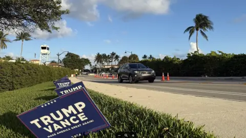 Stephen Greaves/BBC News A car drives past Mar-a-Lago - there are Trump Vance campaign signs in the grass verge and a US Secret Service control tower is seen in the distance