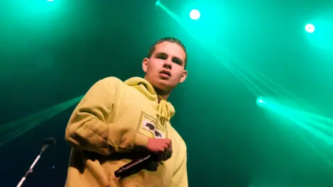 Slowthai wearing a yellow hoodie and holding a black mic. He has short brown hair. There are green lights behind him.