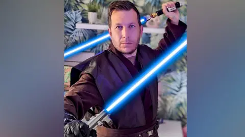 Chris Saunders, dressed in brown Star Wars costume and holding two blue lightsabers, one in each hand. Tropical style, green leaf pattern wallpapers is out of focus behind him.