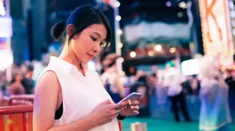 A woman in a white sleeveless top stands in New York City's Times Square at dusk, looking down at the smartphone in her hands