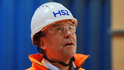 Wearing a white hard hat, safety goggles and an orange high visibility jacket, HS2 Ltd CEO Mark wild appears to be looking uptowards something out of shot as the camera takes his picture