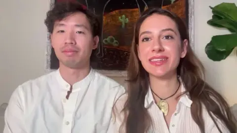 Jeff Tan and Yeganeh Mafaher sit next to each other in front of a white wall with an art piece and a plant.