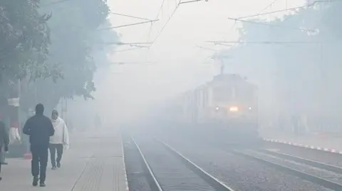 Commuters walking on railway tracks wearing caps and jackets as a train arrives amid fog at Shahabad Mohammadpur railway station in the morning winter chill and fog in New Delhi, India.