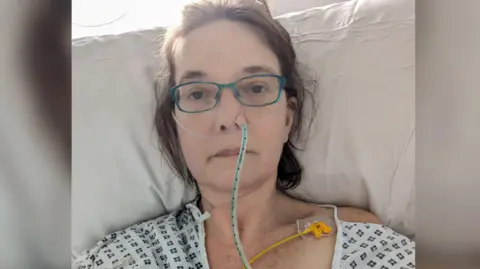 Bells Lewers has a nasogastric tube and an oxygen tube attached to her nose. She also has what appears to be a central line attached to her chest. She has brown hair and turquoise glasses and is lying on a pillow on a hospital bed, wearing a hospital gown.