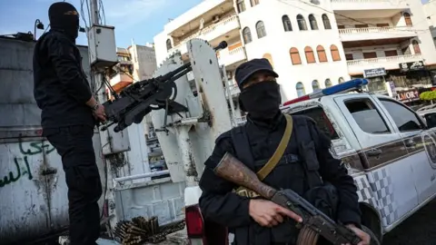 Two Syrian security troops man a checkpoint in Latakia - both carrying machine guns with one stood on the back of a truck