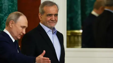 Vladimir Putin, seen in profile, gestures towards his Iranian counterpart, Masoud Pezeshkian, who is walking slightly ahead of him. Both men are wearing dark suits. Putin has a white shirt and dark tie, while Pezeshkian wears a blue open-necked shirt.