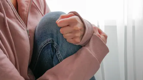 A stock image of a woman kneeling on a chair, wearing a pink jumper and blue jeans, with her hands and arms wrapped around her leg. Her face is not visible.