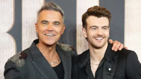 Robbie Williams has a huge smile and his arm around Jonno Davies who is also smiling. Both smartly dressed at a premiere event for Better Man.