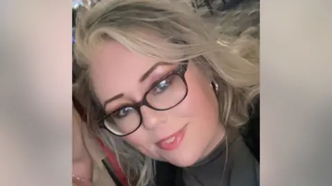 Kimberley Dyson in a cropped selfie image. She is smiling. She has blond hair, brown eyebrows, eyeshadow and dark glasses. She wears a thin gold chain, gold hoop earrings, and a nose ring.