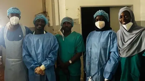 A group of medics at Saudi Hospital in el-Fasher in scrubs smile as they stand for a group photo after successfully performing two-hour emergency caesarean under bombardment. Dust from the shell damage can be seen on the floor