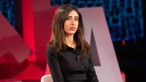 A young woman with long brown hair, dressed in a black shirt, sits on a white chair in a TV studio