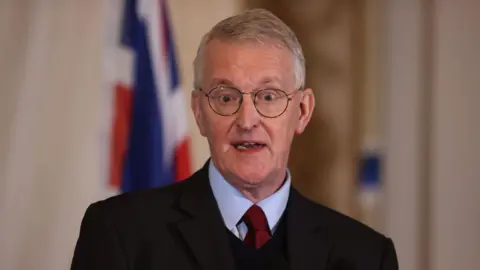 Benn has short hair and is wearing glasses. He is wearing a black suit with a blue shirt and a red tie.