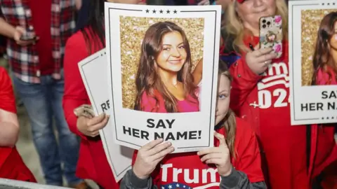 Trump rallygoers hold a sign of Riley's face saying 'say her name'