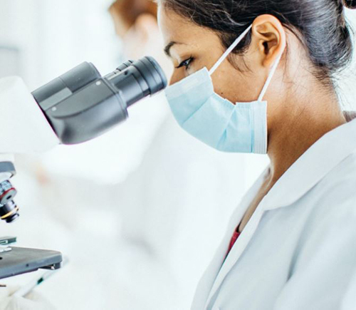 Masked researcher looking into microscope