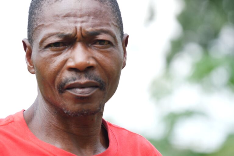 A man in a red t-shirt gazes into the camera, squinting slightly against the light in Budjala territoire, Sud-Ubangi, Democratic Republic of Congo. Image by Francesco De Augustinis for Mongabay.