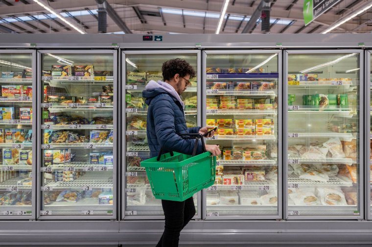 Man shopping in a supermarket while on a budget. He is looking for low prices due to inflation, standing looking at his phone in front of a row of freezers. He is living in the North East of England.