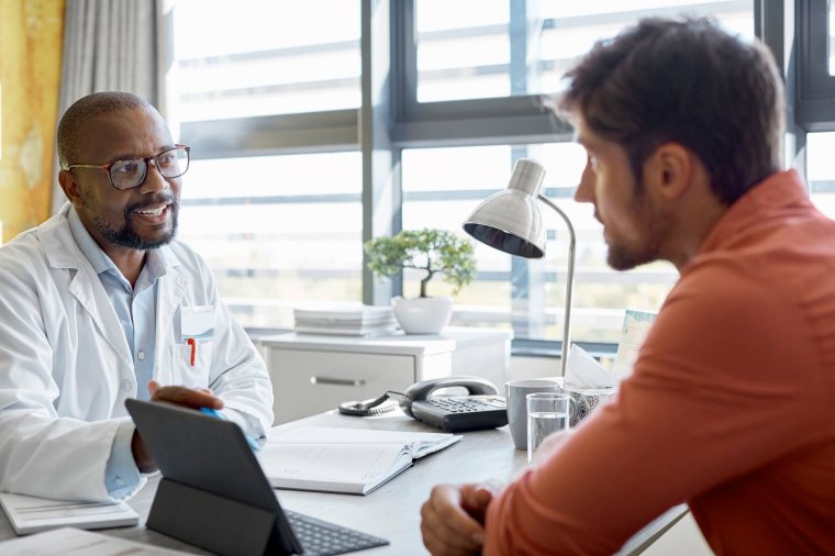 Doctor discussing with man over digital tablet. Young male is visiting healthcare worker in clinic. They are sitting at desk.