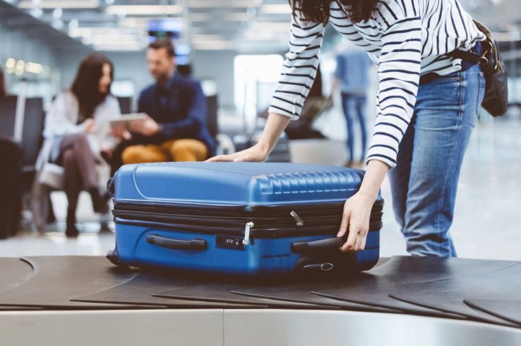 Young woman passenger collecting her luggage from conveyor belt. Female traveler picking up suitcase from baggage claim line in airport terminal.