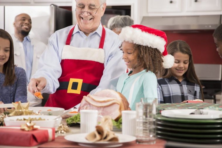 Multi-ethnic group of family members including senior adults, mid-adults, and children all work together in a home kitchen to prepare dinner for family Christmas celebration. Christmas tree, holiday food items, and various decorations in scene.