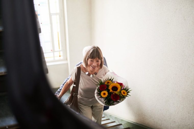 Senior woman walking up the stairs with flowers. Female with husband behind walking up the staircase holding a flower bouquet.