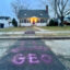 A house with its lights on is in the background. In the foreground: "GEO" spray painted in purple on the sidewalk leading up to it.