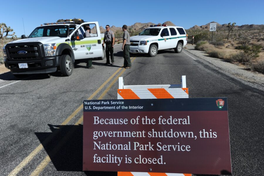 Rangers beside vehicles are in the background. In the foreground, a sign reads: "Because of the federal government shutdown, this National Park Service facility is closed."