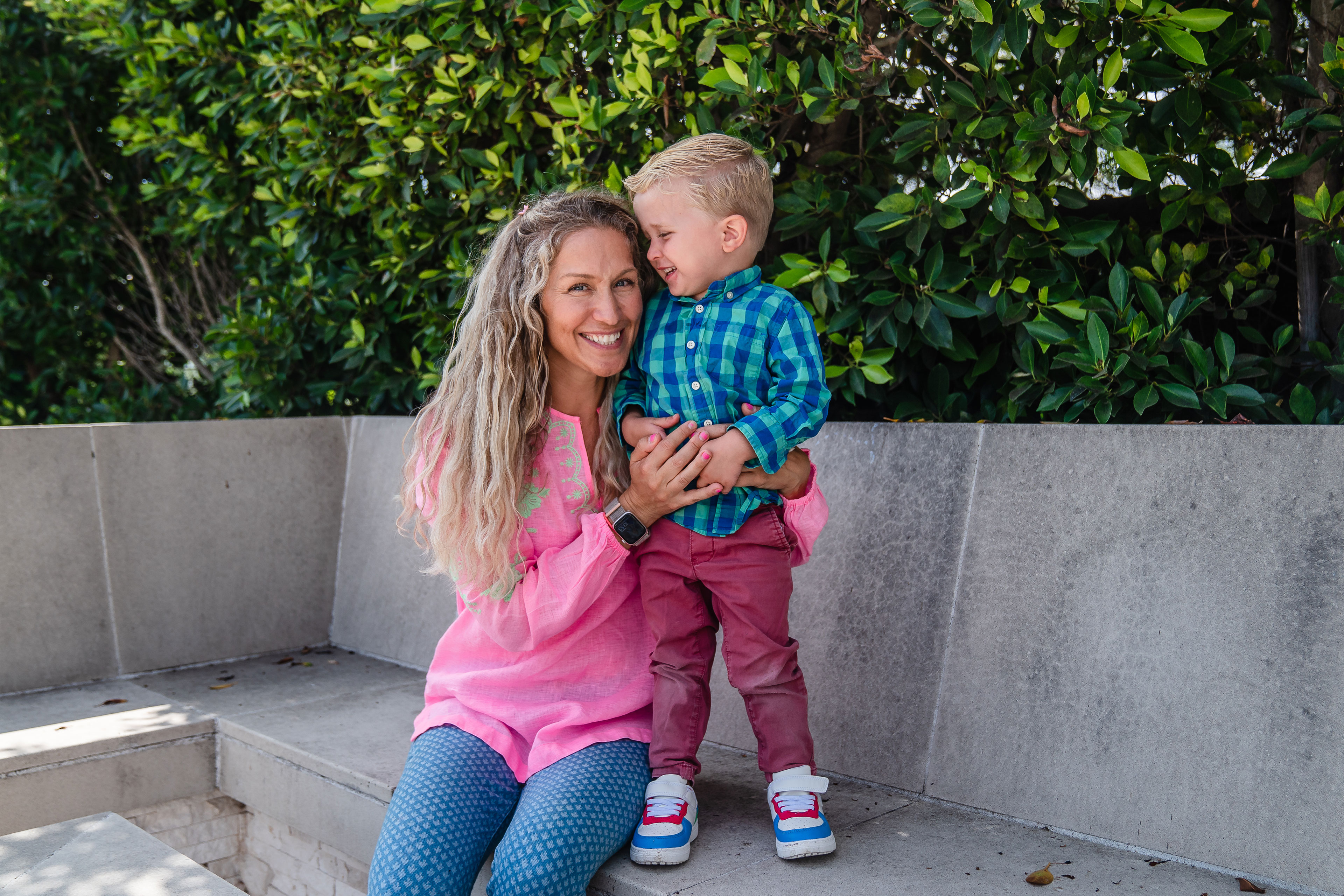 A woman sits on a concrete bench with her arm around a young boy standing on the bench beside her. Both are smiling