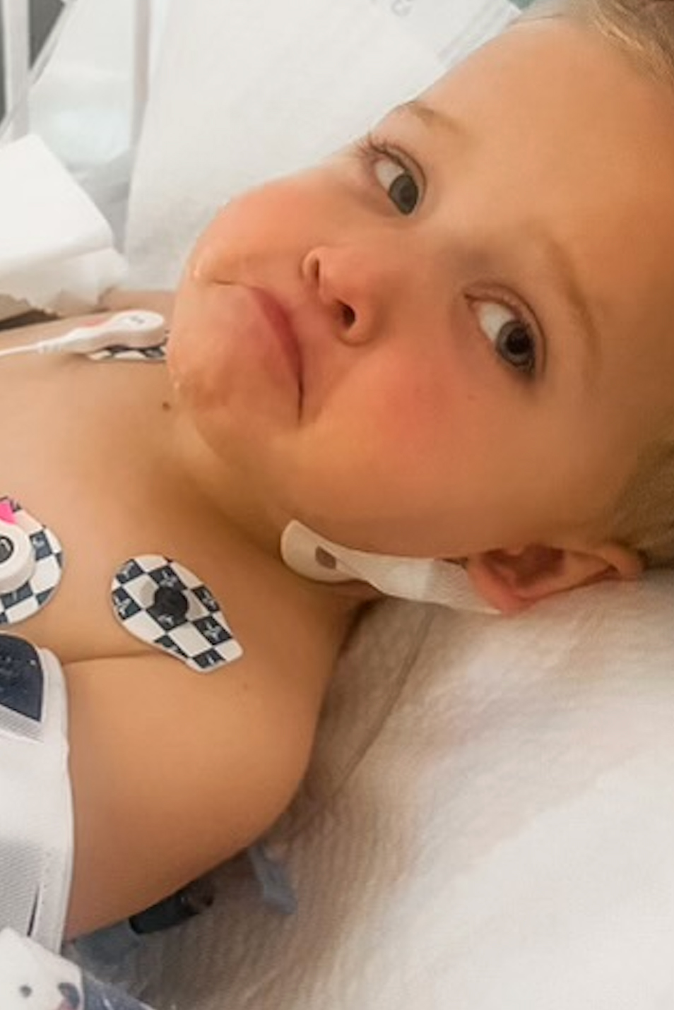 A sad-looking young boy lies in a hospital bed with a bandage on his neck and electrodes on his chest.