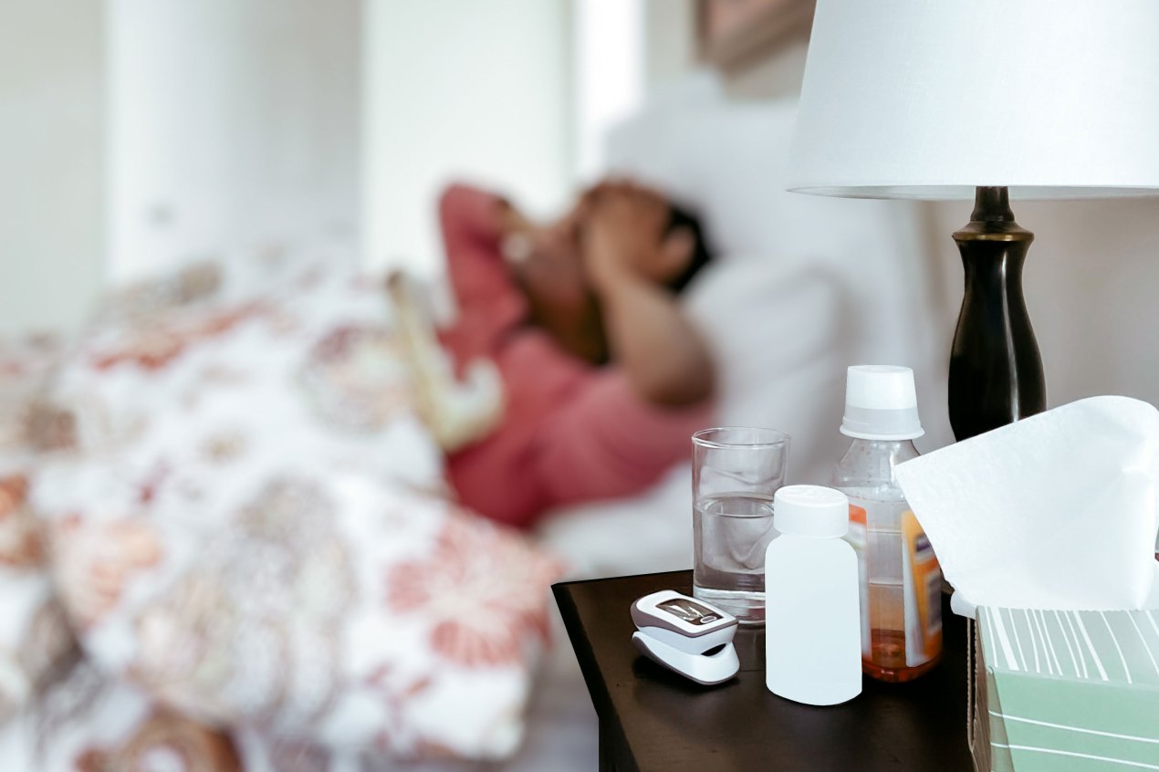 Close-up of cold medication, a pill bottle, a glass of water, a pulse oximeter, and a box of tissues on nightstand with a person lying in bed with hands on their head out-of-focus in the background.