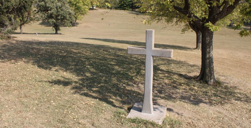 A photograph showing a stone, weathered, 6-foot-tall cross in a graveyard on a sunny day.