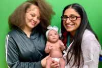 Two women smile at the camera while holding a baby with a white bow on her head