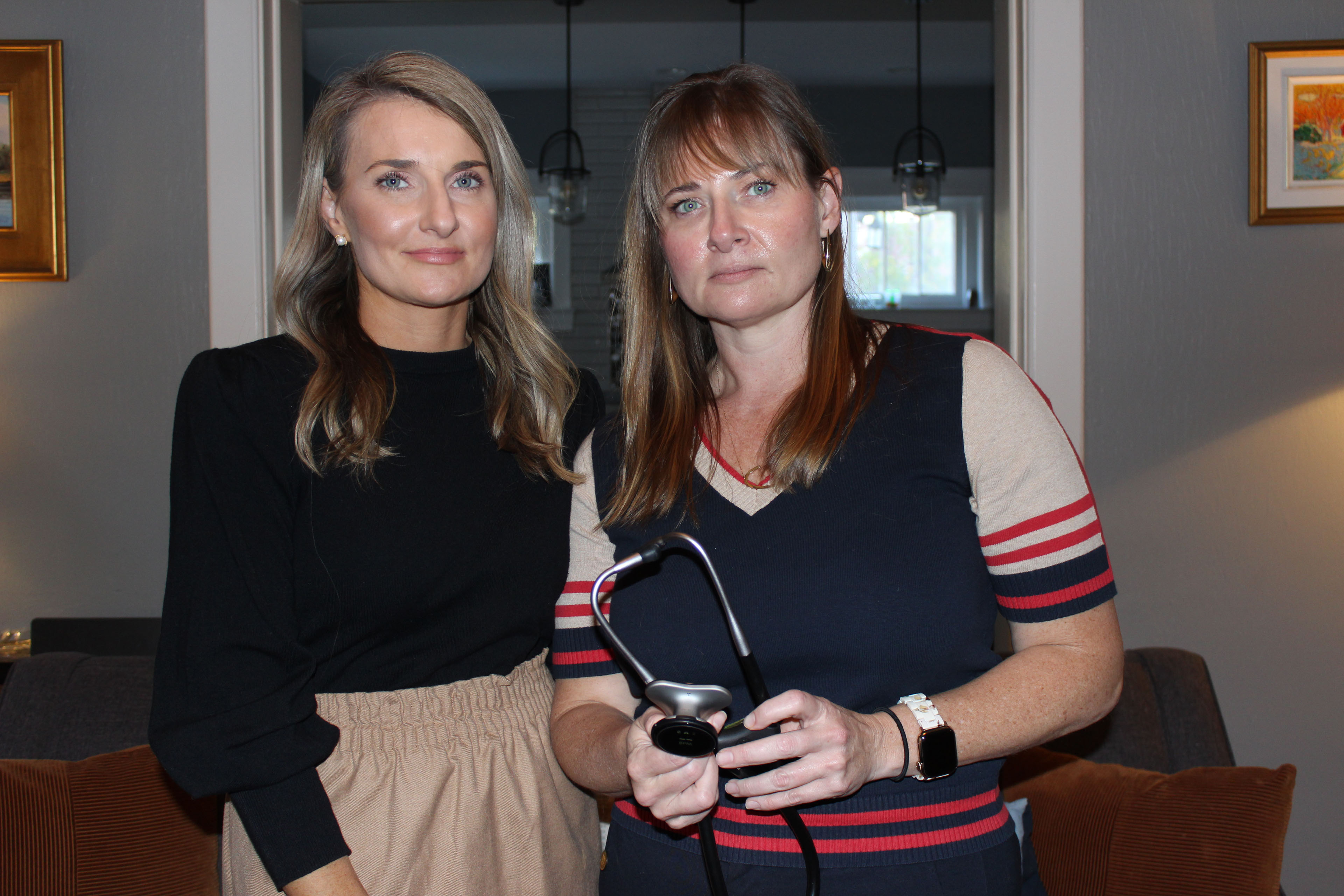 Two adult sisters stand side by side in their home. The woman on the right holds a stethoscope in her hands.