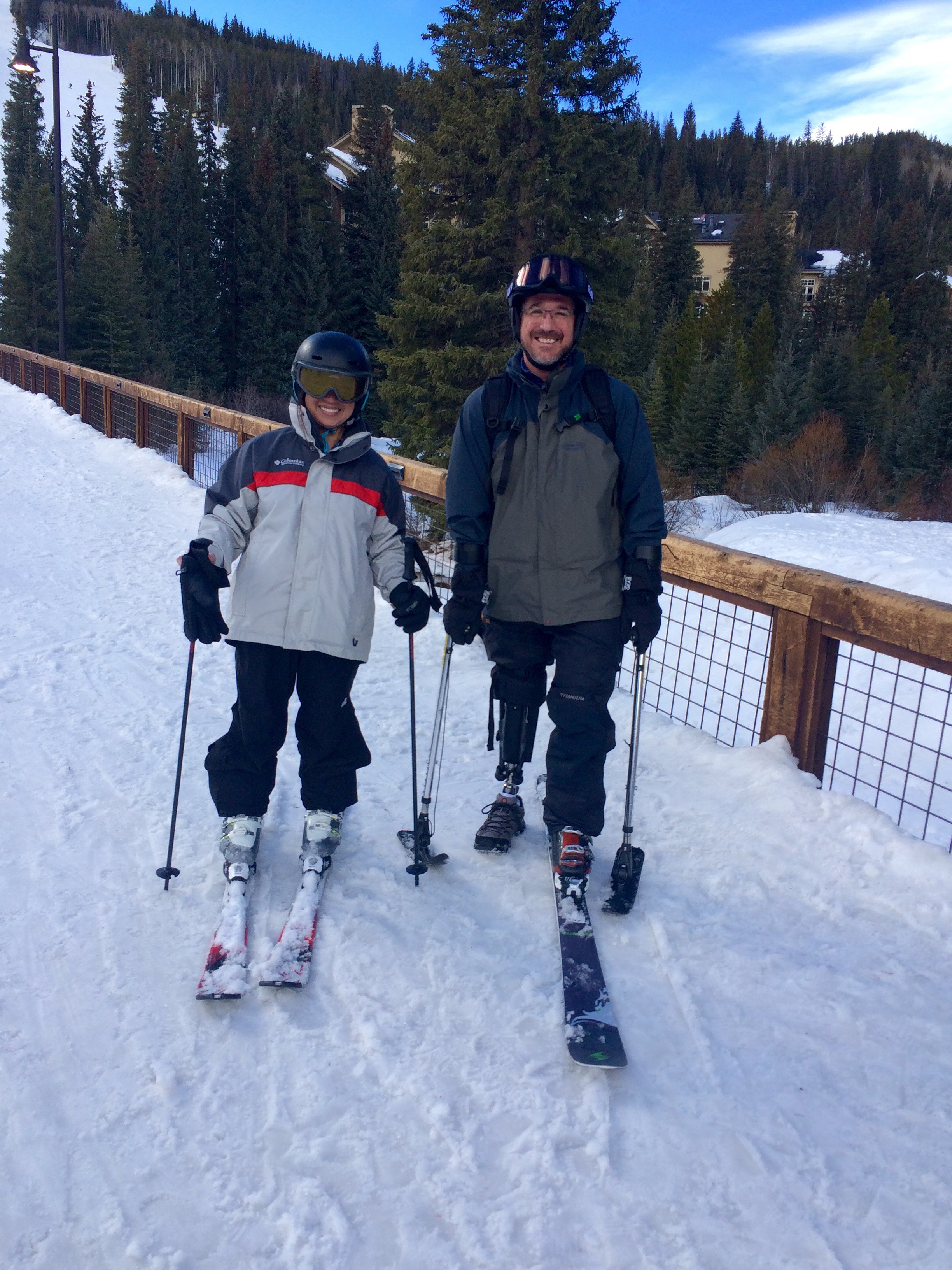 A man with a prosthetic leg stands beside a woman. They are on a snowy ski slope and dressed in snow gear.