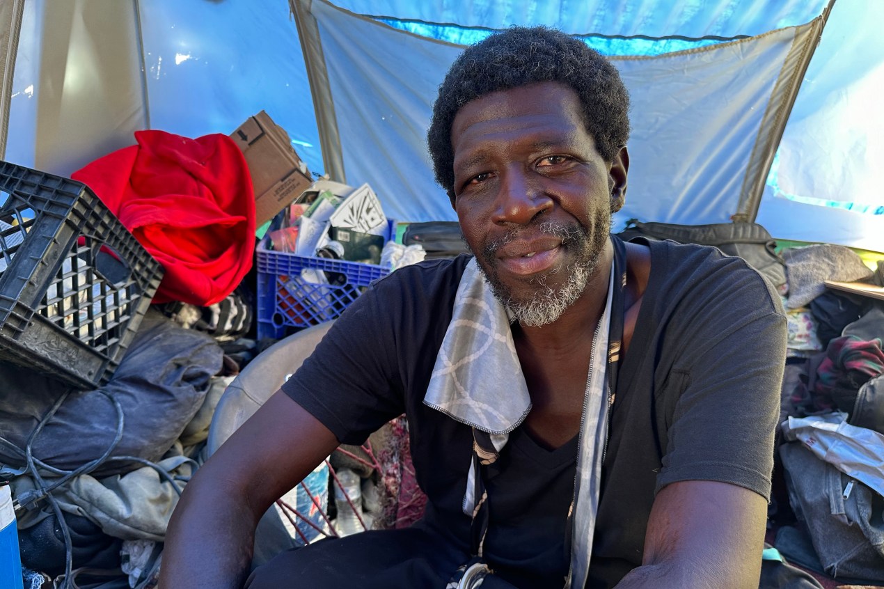 A portrait of Maurice Clark, who is sitting amongst his belongings. He has a warm expression as he looks towards the camera.