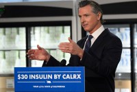 California governor Gavin Newsom stands behind a blue and white sign that reads "$30 insulin by CalRx"