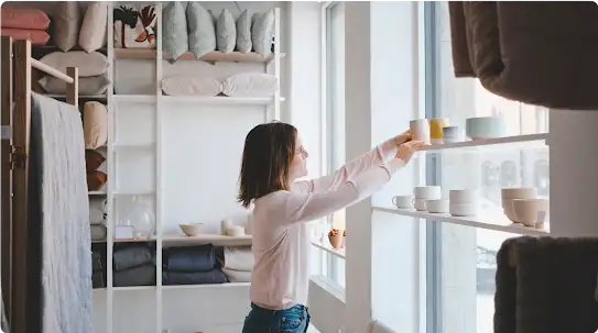 Une propriétaire d’entreprise range des produits sur une étagère, en vitrine de son magasin d’articles de maison.