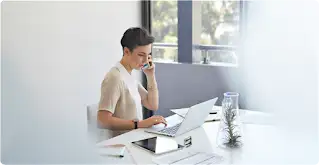 Une femme aux cheveux courts est assise à son bureau, sur lequel sont posés un ordinateur portable et une tablette. Elle parle au téléphone à l’aide d’un téléphone intelligent.