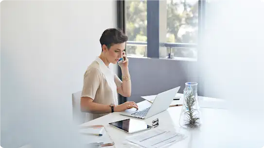 Uma mulher de cabelos curtos sentada em uma mesa e conversando em um smartphone com um laptop e um tablet na frente.