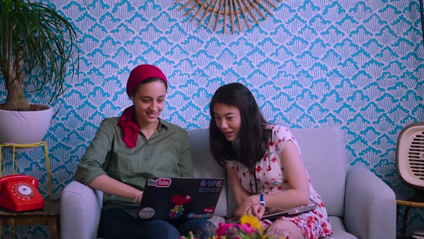 Two women Googlers of Middle Eastern and Asian backgrounds sit together on a couch, reviewing work on a laptop