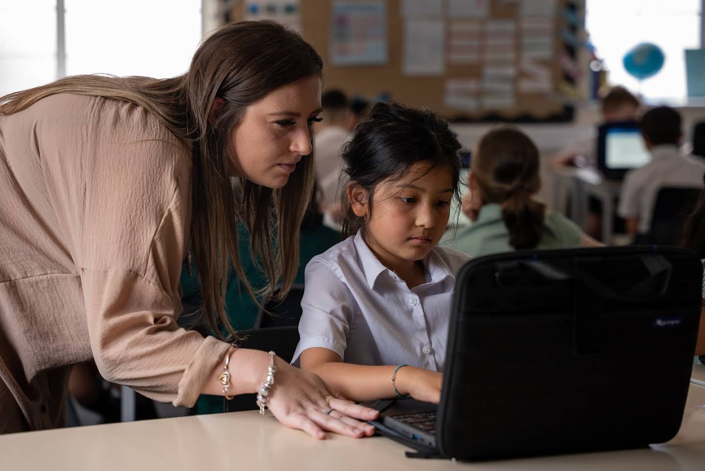 A teacher in classroom looks over a young student’s work on a laptop.