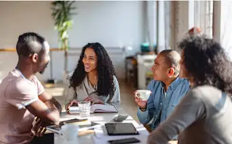 Four team members sit at a table having a coffee-work meeting with notebooks, tablets and phones across the table.