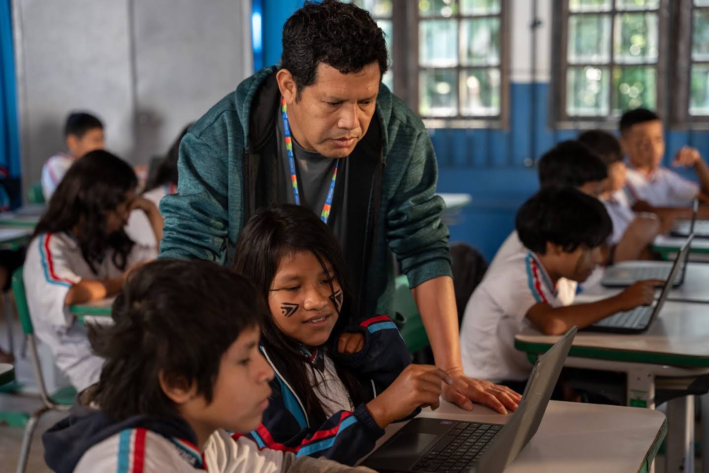 A teacher in a classroom checks two students’ work on a laptop.