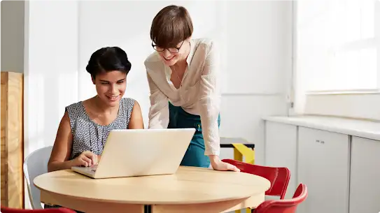 Deux femmes souriantes travaillent ensemble sur un ordinateur portable autour d’une petite table ronde. L’une d’elles porte un haut à motifs, et l’autre, un haut blanc.