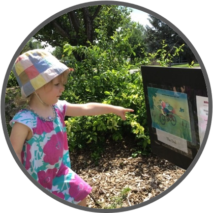 two-year-old girl in dress and sun hat points at a storybook page posted on a sign outdoors