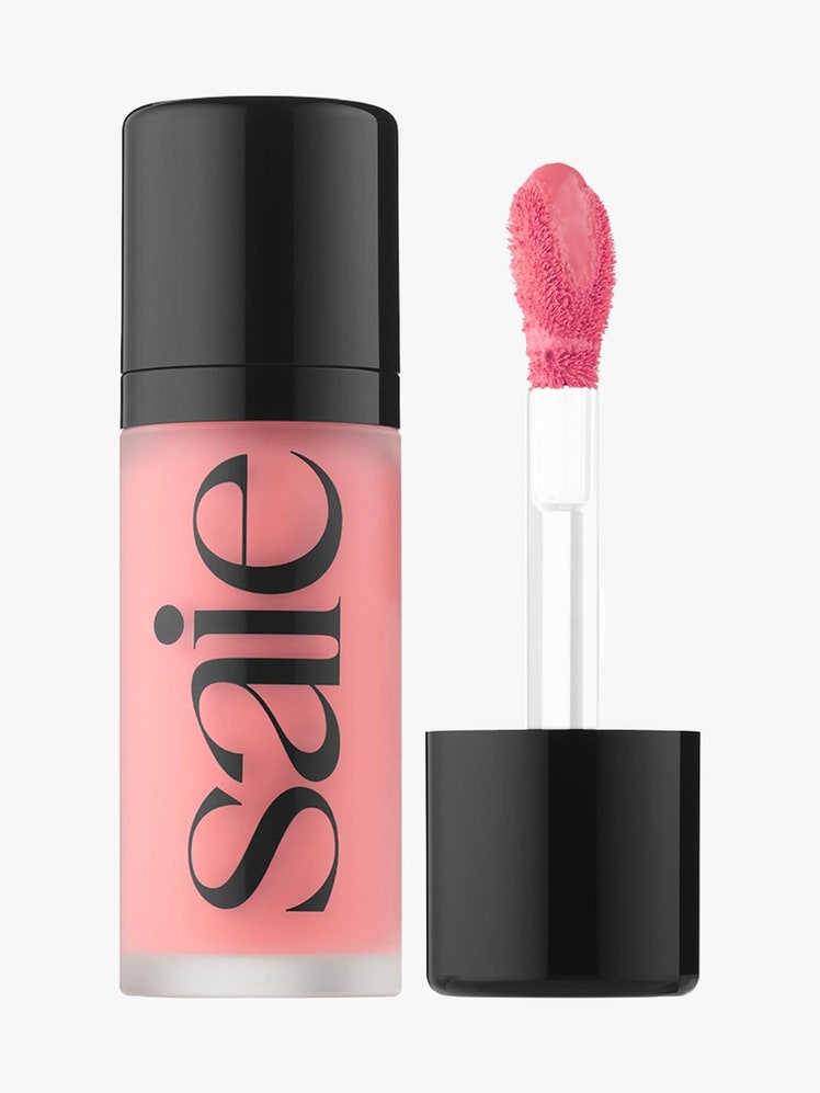 Saie Dew Blush Liquid Cheek Blush: A clear tube of liquid pink blush with black cap and text alongside its matching applicator on a light gray background