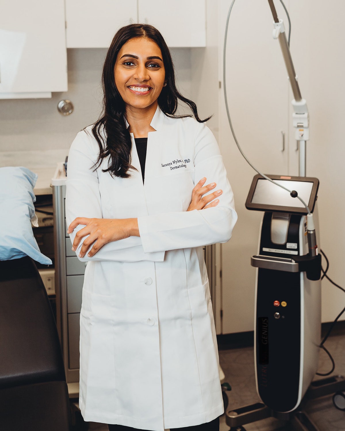 Dermatologist Saranya Wyles shown standing next to a device in her dermatology practice wearing a lab coat