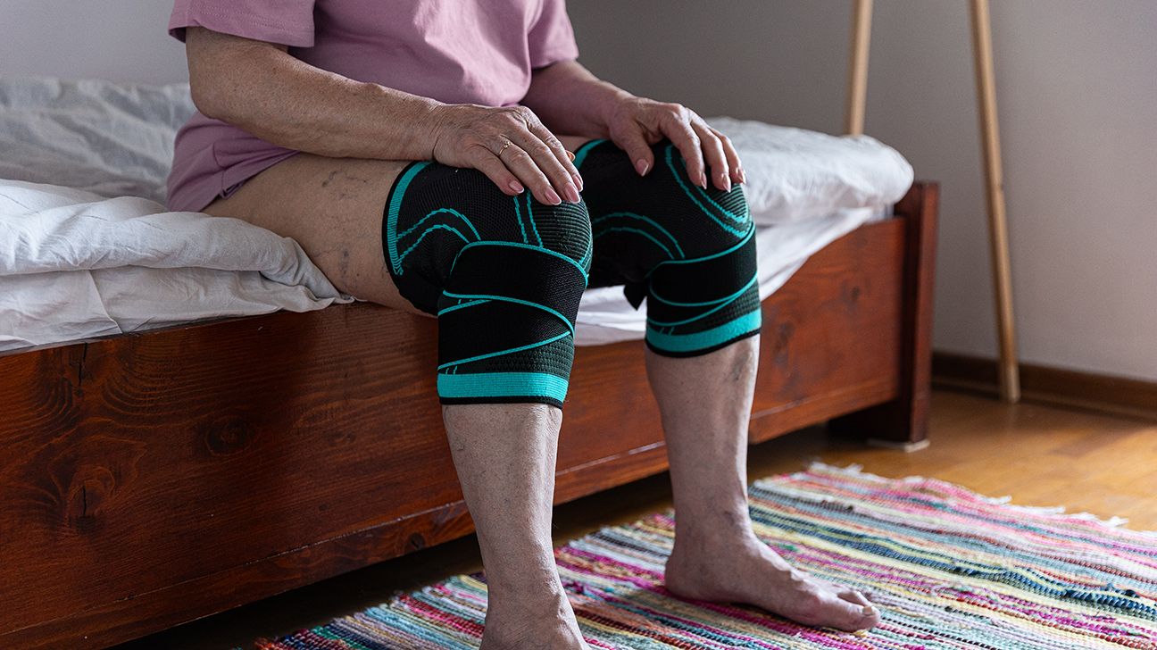 Older adult sitting on a bed with two knee braces for osteoarthritis